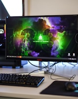 A gaming setup featuring a central monitor with a vibrant display showing 'The Spirit of Gaming'. On either side, there are gaming PCs with illuminated components and transparent cases. Headsets hang from the side of the monitor. The desk also has a black keyboard and mouse, and a branded mousepad.