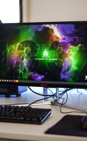 A gaming setup featuring a central monitor with a vibrant display showing 'The Spirit of Gaming'. On either side, there are gaming PCs with illuminated components and transparent cases. Headsets hang from the side of the monitor. The desk also has a black keyboard and mouse, and a branded mousepad.