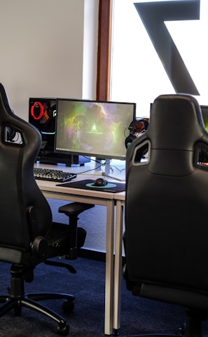 A row of gaming stations equipped with large monitors, gaming chairs, keyboards, and headphones. The setup is organized and ready for gaming, featuring a colorful screen display. The room has a modern and tech-savvy ambiance.