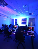 A modern gaming setup with multiple high-backed gaming chairs and monitors arranged in a room with a cool blue lighting. The computers have illuminated keyboards and glowing screens, creating a high-tech ambiance.