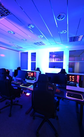 A modern gaming setup with multiple high-backed gaming chairs and monitors arranged in a room with a cool blue lighting. The computers have illuminated keyboards and glowing screens, creating a high-tech ambiance.