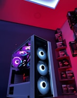 A gaming computer setup featuring a high-performance PC tower with multiple RGB fans lit in blue and purple hues. The case has a modern design with transparent panels showcasing internal components. In the background, shelves are stocked with various Ryzen and other gaming components under a vibrant red and blue ambient light.
