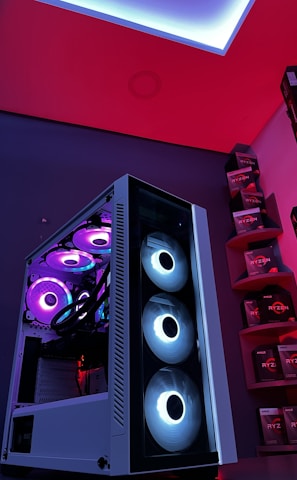 A gaming computer setup featuring a high-performance PC tower with multiple RGB fans lit in blue and purple hues. The case has a modern design with transparent panels showcasing internal components. In the background, shelves are stocked with various Ryzen and other gaming components under a vibrant red and blue ambient light.