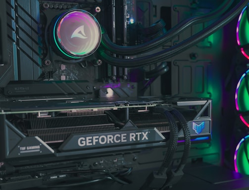 The image depicts the inside of a computer case featuring a high-performance graphics card labeled 'GEFORCE RTX' with RGB lighting. The cooling fans are illuminated with vibrant colors, creating a visually striking look. Various cables and components are neatly arranged, highlighting advanced hardware and a focus on aesthetics.